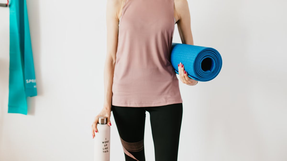 Faceless slim female athlete in sportswear standing with blue fitness mat and water bottle while preparing for indoors workout